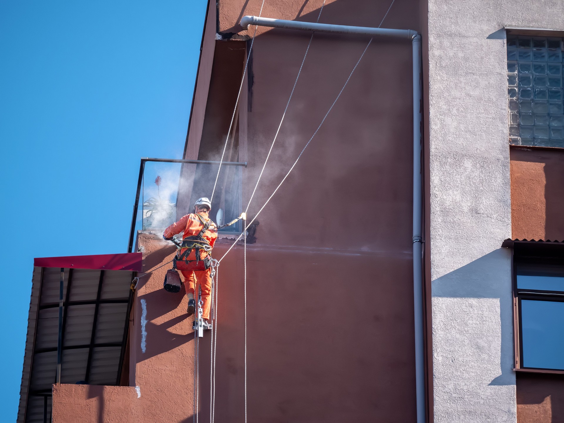 Worker on rope with safety gear performing industrial facade cleaning on tall building. Smoke, dust and tools emphasize action and precision in sunny outdoor environment